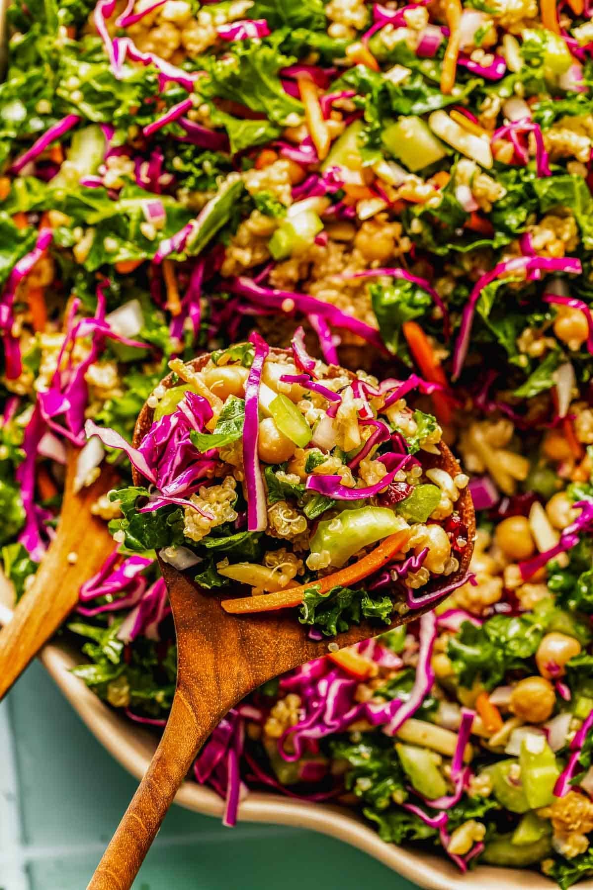 A wooden spoon holds a colorful salad with shredded kale, red cabbage, carrots, cucumbers, chickpeas, and quinoa.