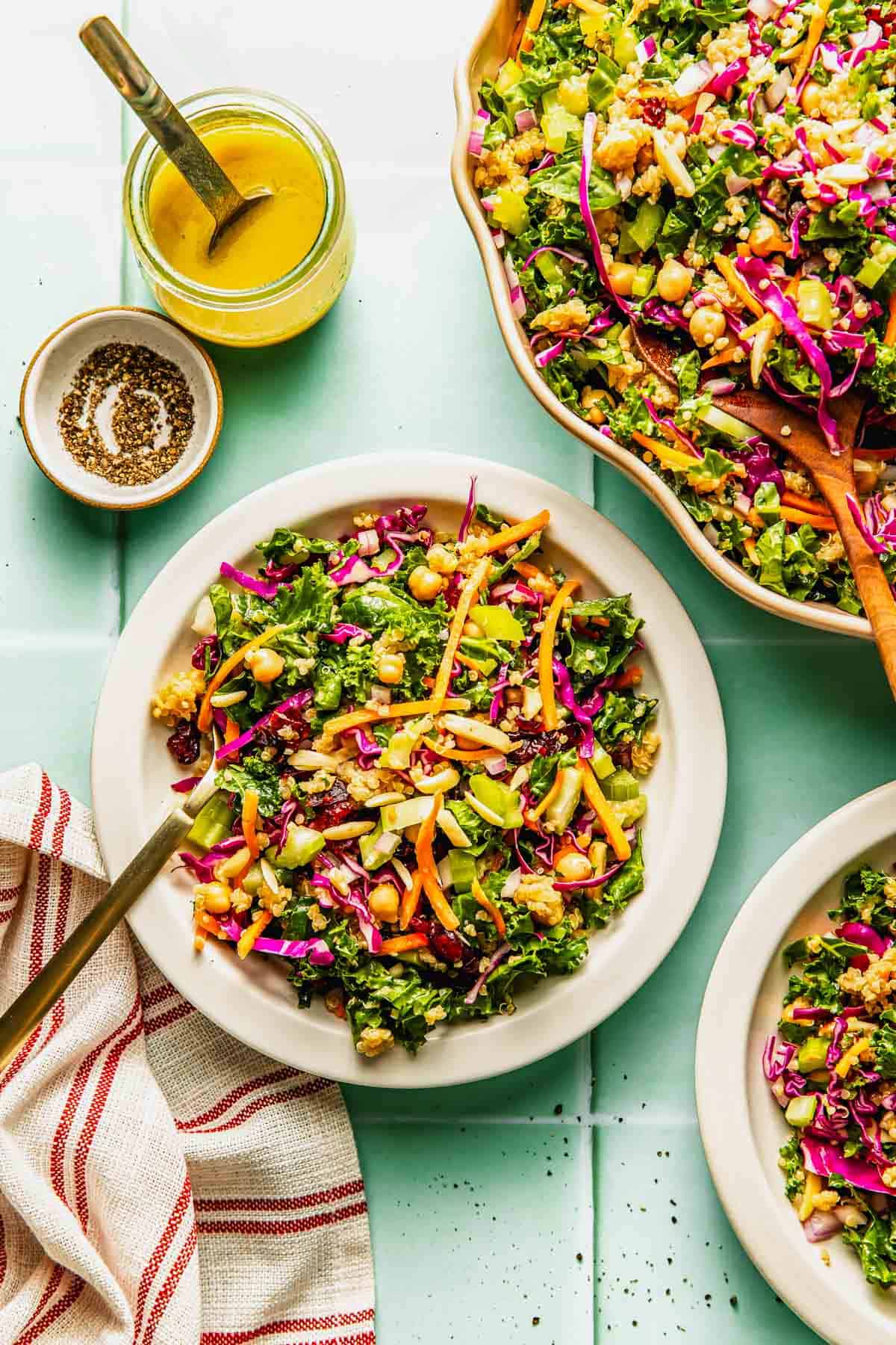 A plate of chopped kale salad with colorful vegetables, chickpeas, and a side of yellow dressing, shown on a light blue table with a striped napkin.