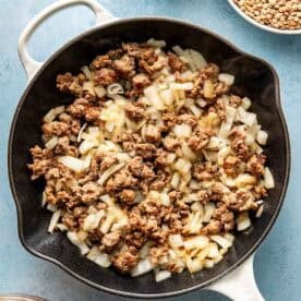 A skillet with cooked sausage and chopped onions, surrounded by bowls of diced sweet potatoes, sliced celery, and dry lentils on a blue surface.