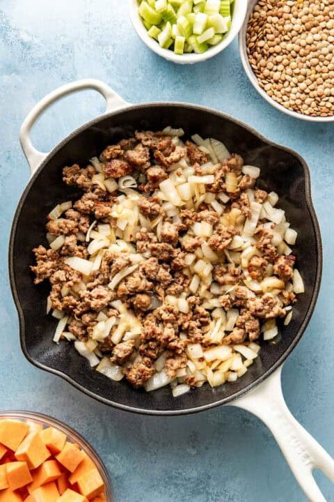 A skillet with cooked sausage and chopped onions, surrounded by bowls of diced sweet potatoes, sliced celery, and dry lentils on a blue surface.