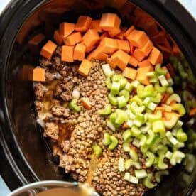 Diced sweet potatoes, celery, ground meat, and lentils in a slow cooker, with broth being poured in.