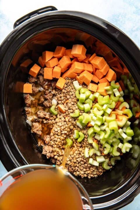 Diced sweet potatoes, celery, ground meat, and lentils in a slow cooker, with broth being poured in.