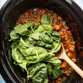 A slow cooker filled with lentil stew, sweet potatoes, and fresh spinach, with a wooden spoon resting inside.