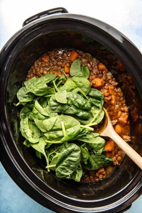 A slow cooker filled with lentil stew, sweet potatoes, and fresh spinach, with a wooden spoon resting inside.