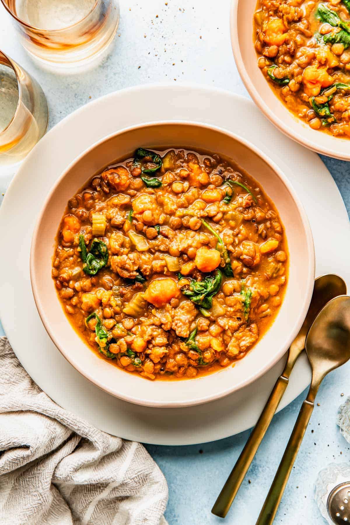 A bowl of lentil soup with vegetables and greens, placed on a white plate with a spoon and fork beside it. A napkin and glasses are partially visible nearby.