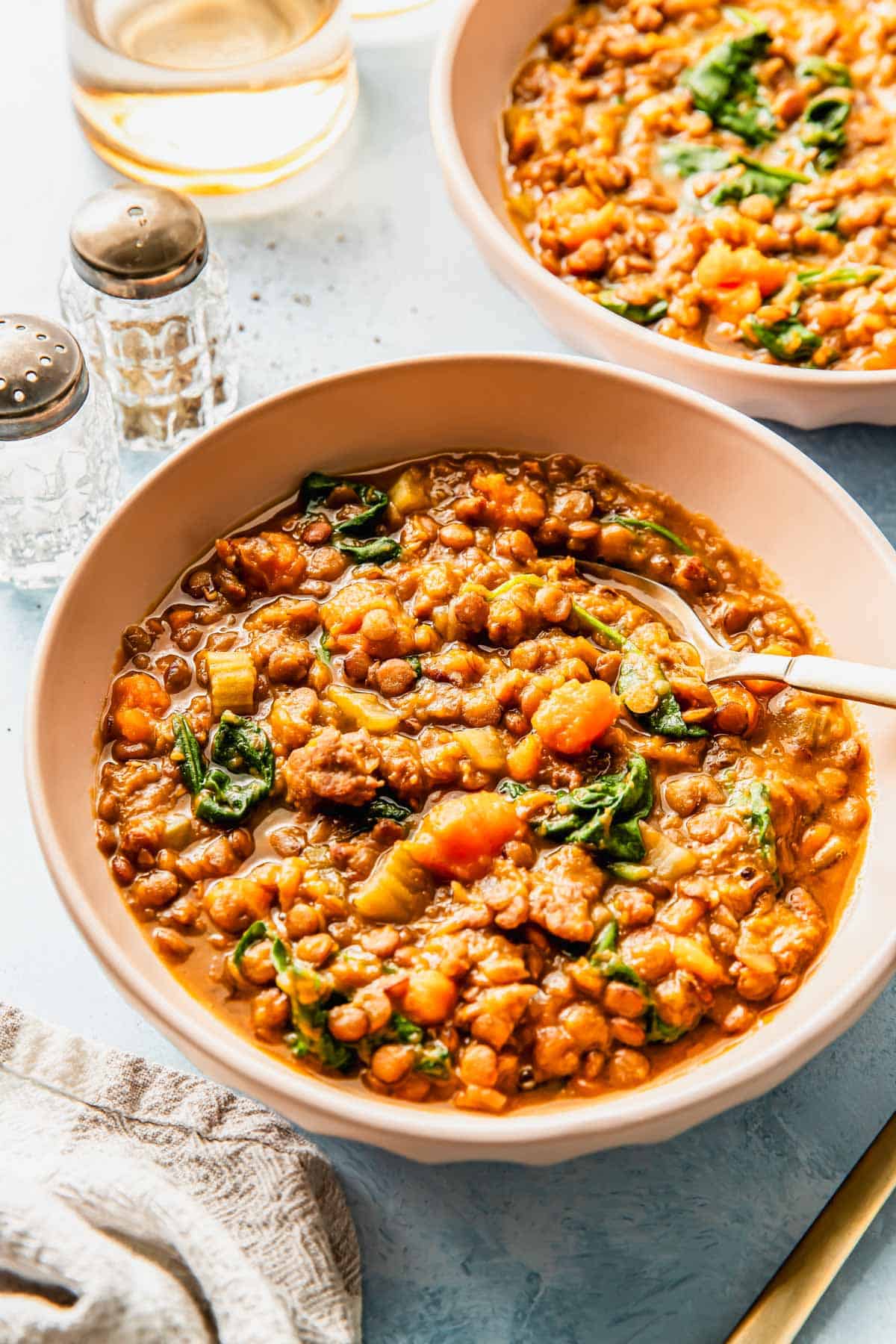 Two bowls of lentil soup with visible spinach, carrots, and celery, served with a spoon, next to a glass of water and salt and pepper shakers on a light surface.
