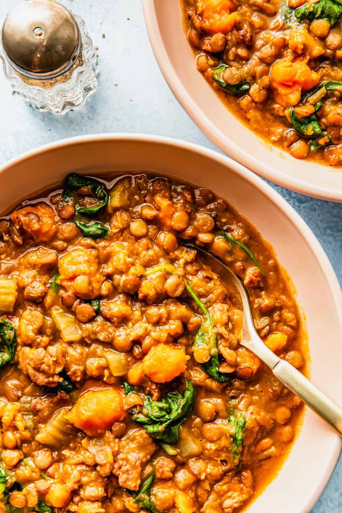 Close-up of two bowls filled with lentil stew containing carrots, spinach, celery, and a golden broth, with a spoon in one bowl. A salt shaker is visible in the background.