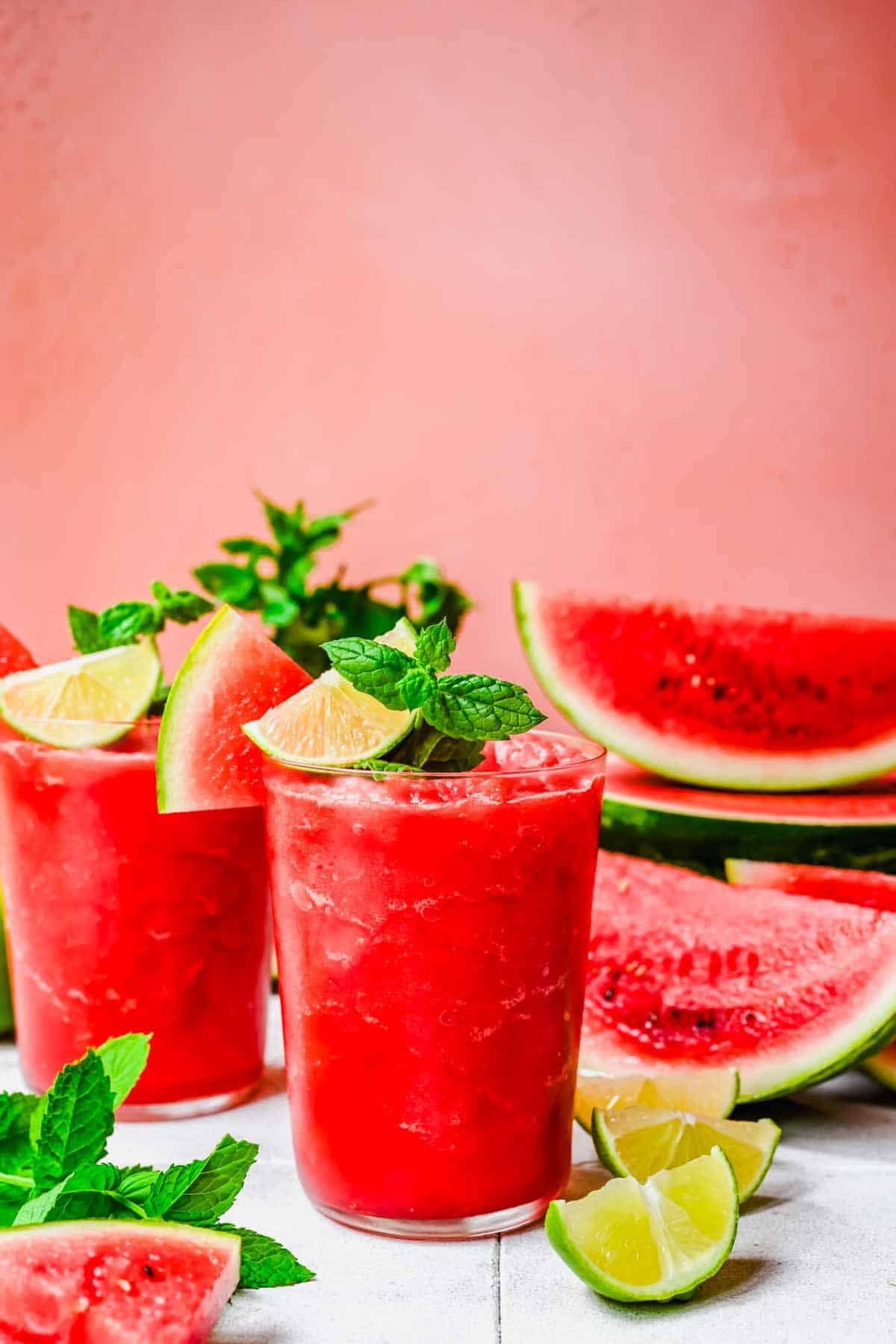 Two glasses of watermelon slushies garnished with lime wedges and mint, surrounded by watermelon slices, lime pieces, and mint leaves on a white surface against a pink background.