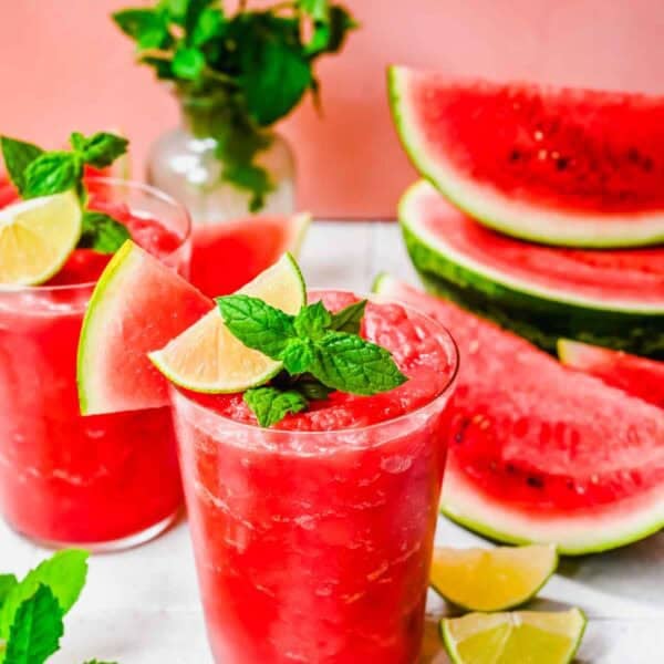 Two glasses of watermelon slushies with ice, lime wedges, and mint garnish. Sliced watermelon and lime, plus a vase of mint, are in the background on a white surface.