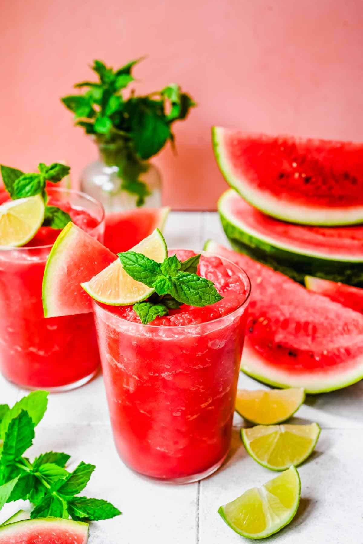 Two glasses of watermelon slushies with ice, lime wedges, and mint garnish. Sliced watermelon and lime, plus a vase of mint, are in the background on a white surface.