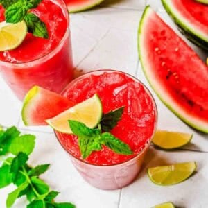 Two glasses of watermelon slushies garnished with lime, mint, and watermelon slice, surrounded by watermelon wedges, lime wedges, and mint leaves on a white surface.