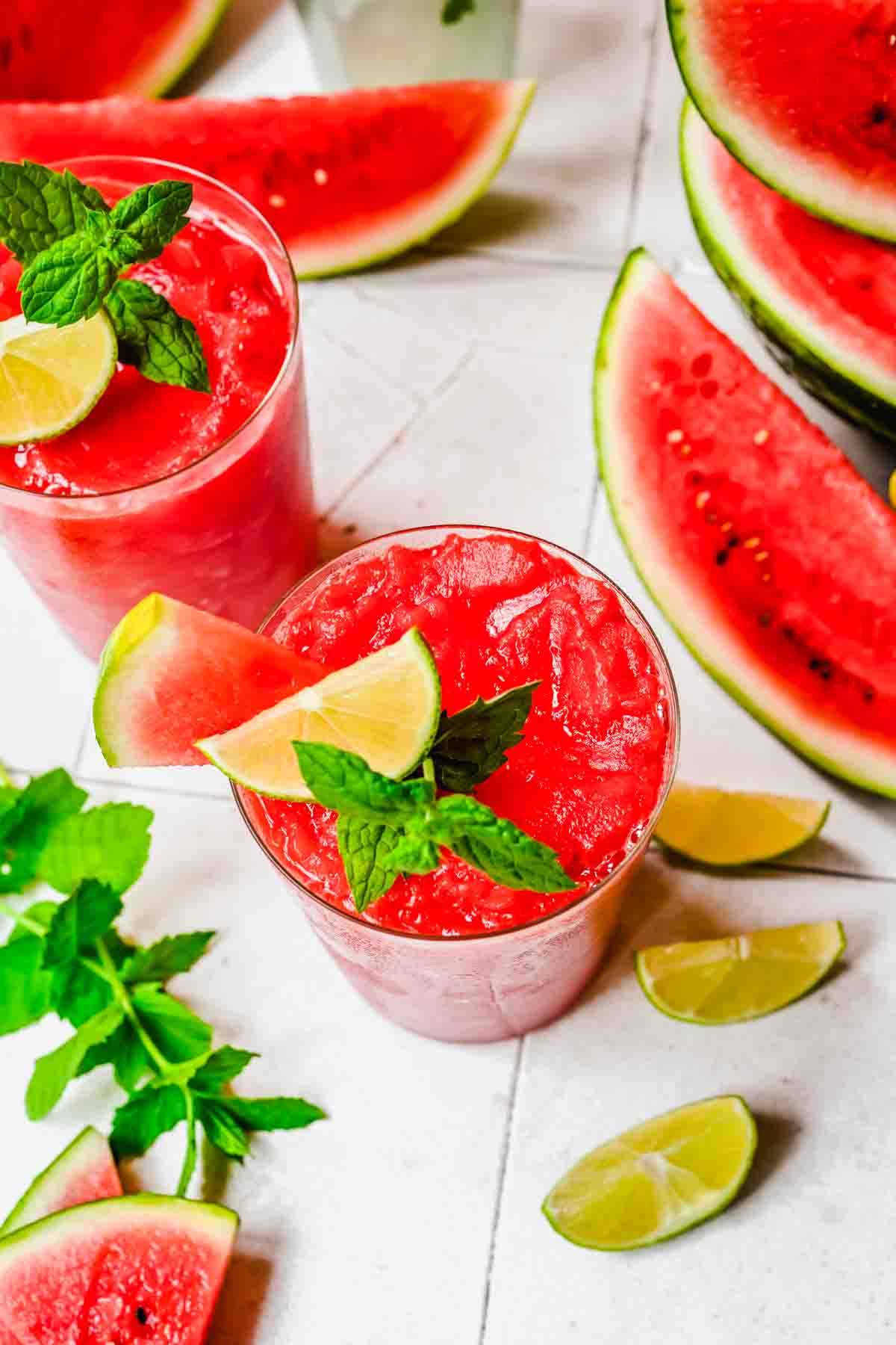 Two glasses of watermelon slushies garnished with lime, mint, and watermelon slice, surrounded by watermelon wedges, lime wedges, and mint leaves on a white surface.