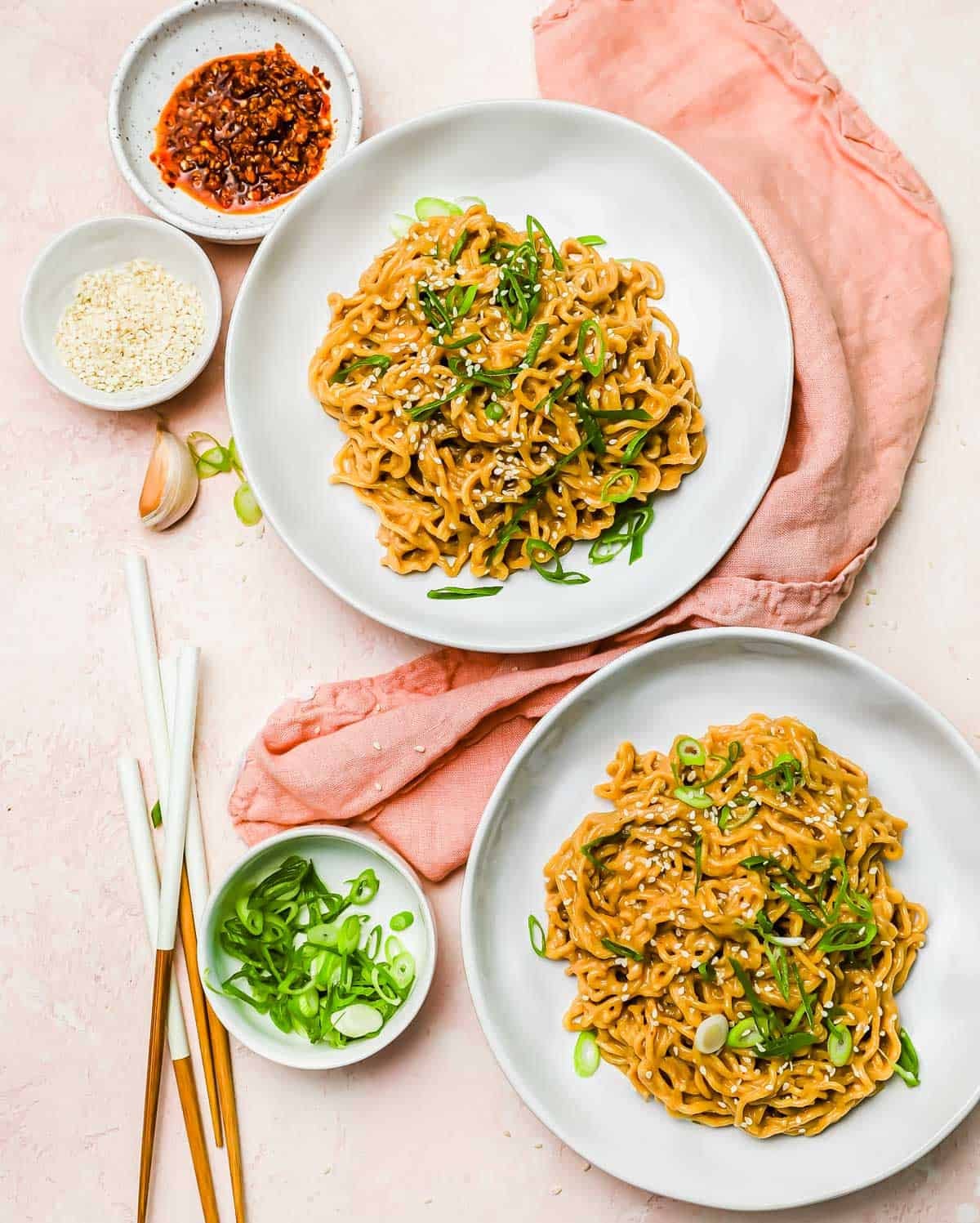 Two plates of saucy noodles topped with sesame seeds and sliced green onions, accompanied by small bowls of chili oil, sesame seeds, and green onions, with chopsticks and a pink napkin.