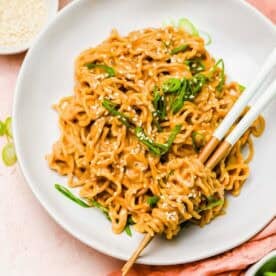 A plate of cooked noodles garnished with sesame seeds and green onions, with chopsticks picking up a portion; bowls of chili oil and sesame seeds are nearby.