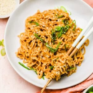 A plate of cooked noodles garnished with sesame seeds and green onions, with chopsticks picking up a portion; bowls of chili oil and sesame seeds are nearby.