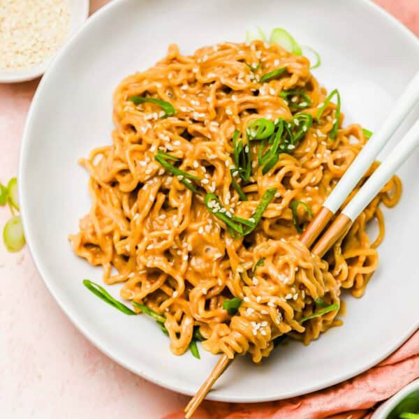 A plate of cooked noodles garnished with sesame seeds and green onions, with chopsticks picking up a portion; bowls of chili oil and sesame seeds are nearby.