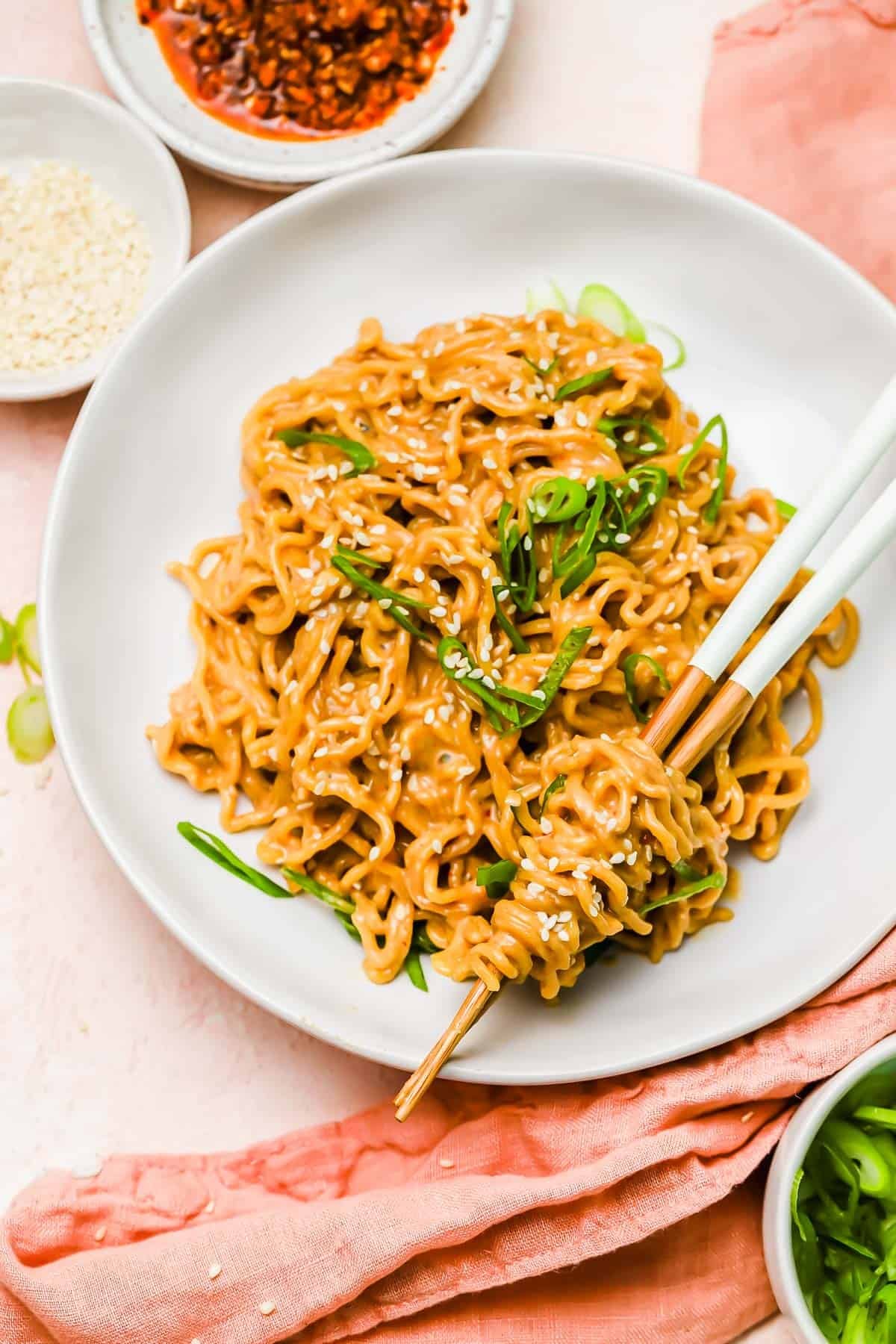 A plate of cooked noodles garnished with sesame seeds and green onions, with chopsticks picking up a portion; bowls of chili oil and sesame seeds are nearby.