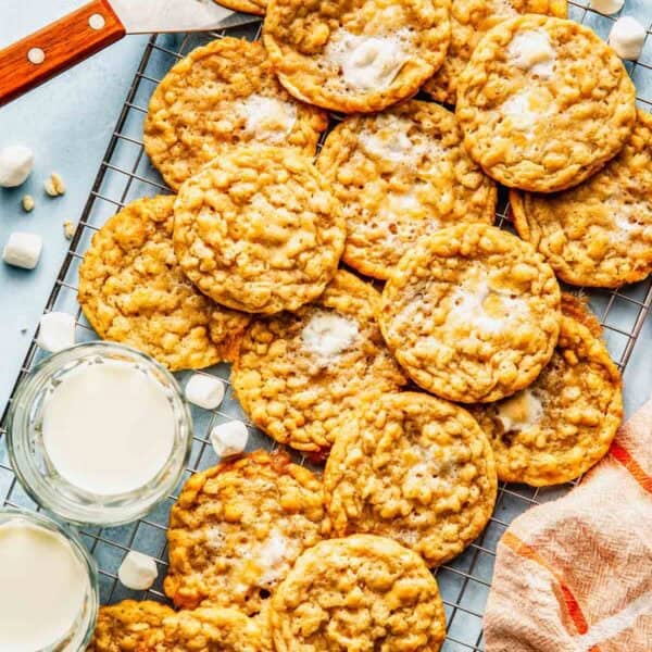 A cooling rack with oatmeal cookies, some topped with melted marshmallows, next to two glasses of milk and a striped towel.
