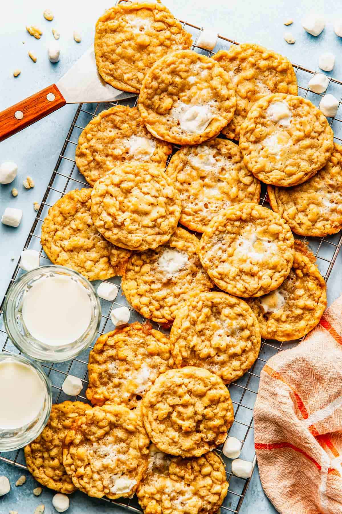 A cooling rack with oatmeal cookies, some topped with melted marshmallows, next to two glasses of milk and a striped towel.