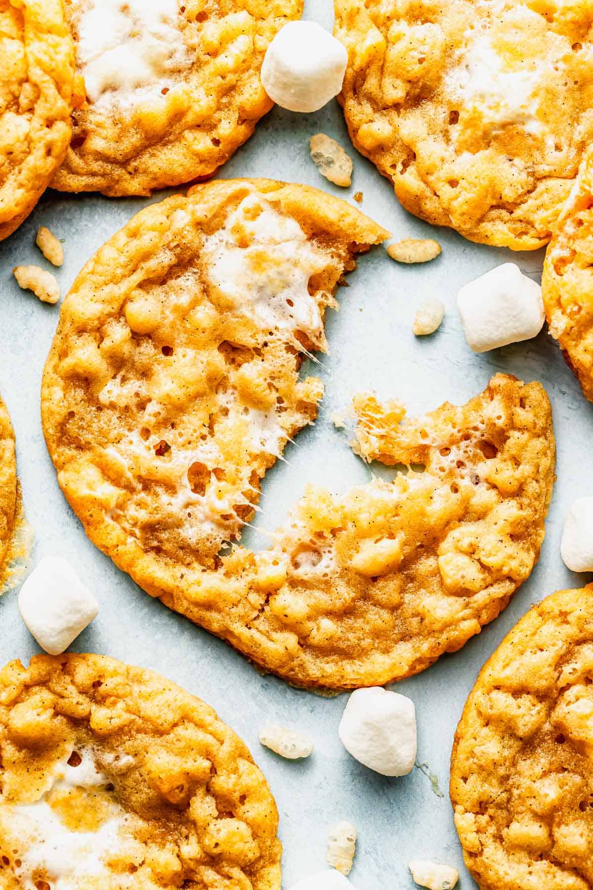A close-up of crispy cookies with bits of marshmallow and rice cereal, one cookie partially eaten, surrounded by small marshmallows.