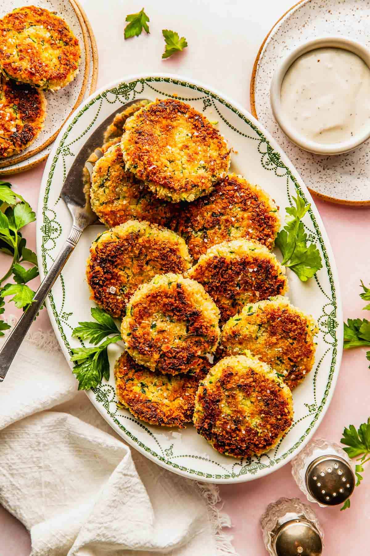 Oval platter with golden-brown patties garnished with parsley, beside a bowl of white dipping sauce, plates, napkin, and salt and pepper shakers on a light surface.