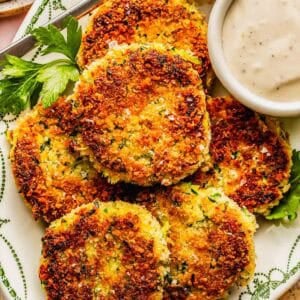 A plate of golden-brown, crispy vegetable patties garnished with parsley, served with a small bowl of creamy dipping sauce.