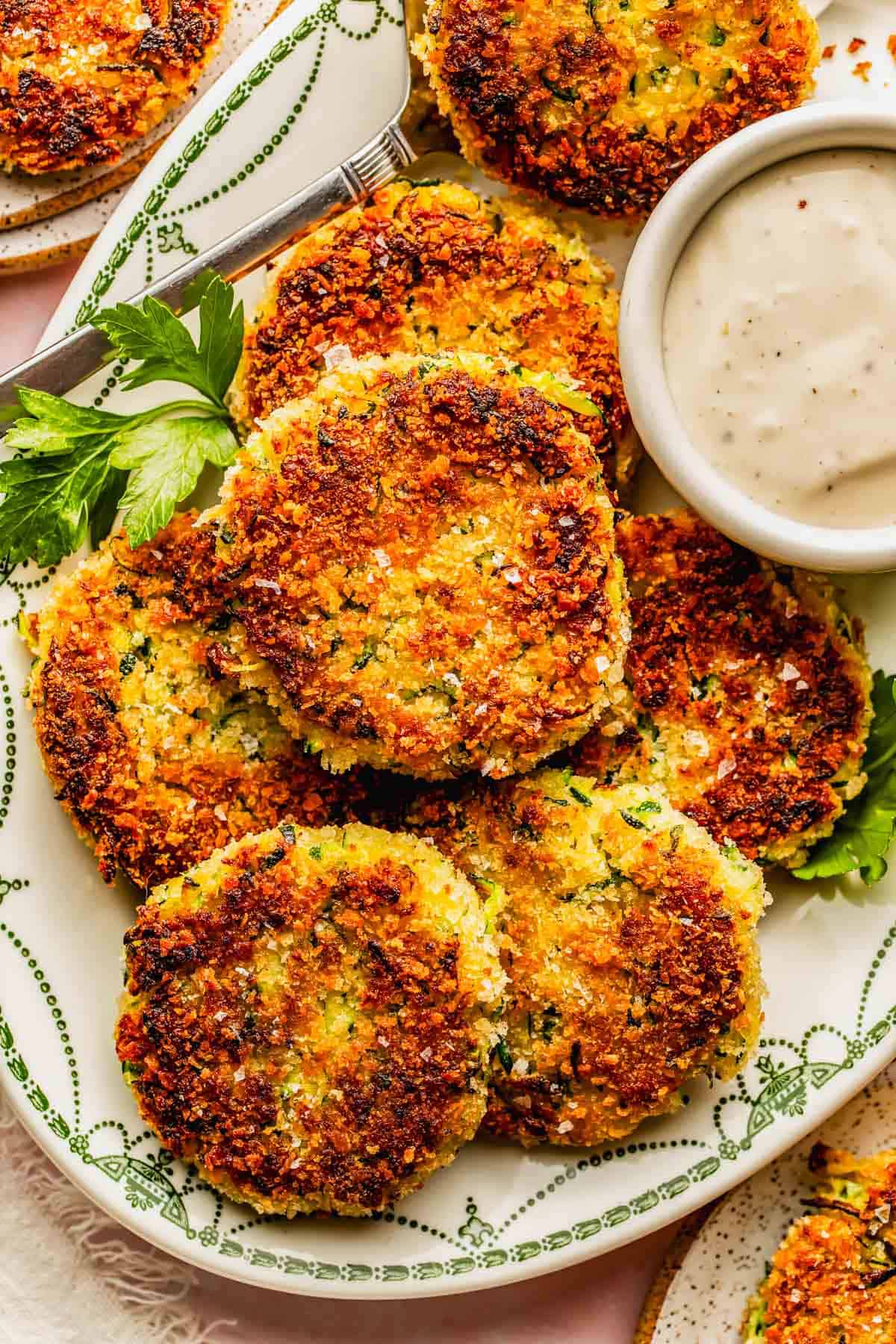 A plate of golden-brown, crispy vegetable patties garnished with parsley, served with a small bowl of creamy dipping sauce.