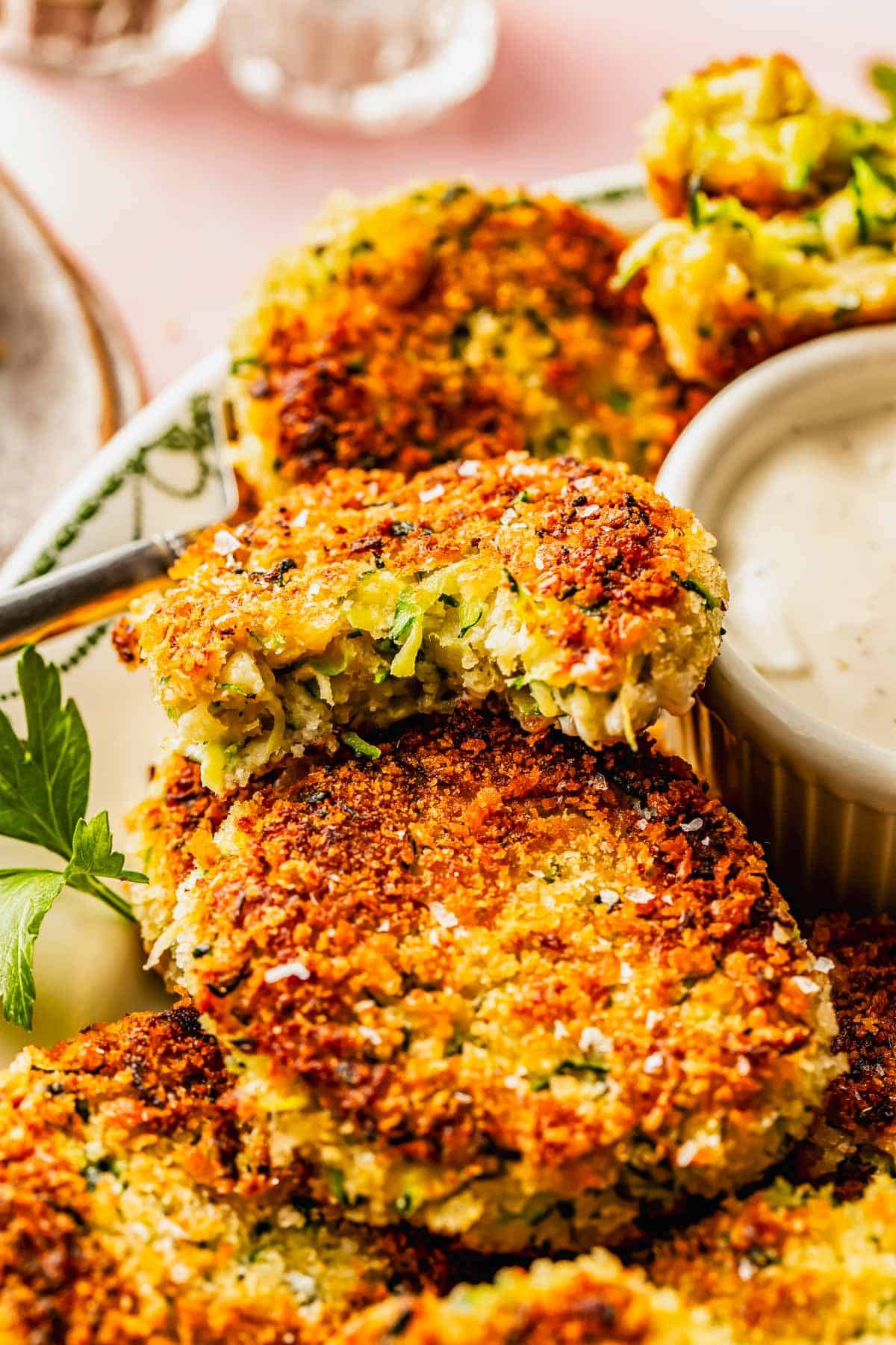 Golden-brown zucchini fritters stacked on a plate with a small bowl of creamy dipping sauce and a sprig of parsley.