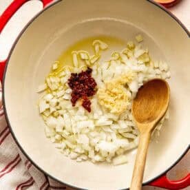 A red Dutch oven containing diced onions, minced garlic, and sun-dried tomatoes in oil, with a wooden spoon and small bowls of salt and pepper nearby.