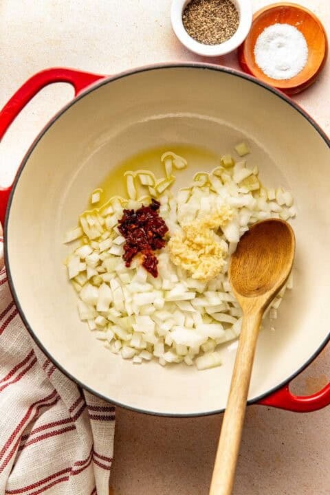 A red Dutch oven containing diced onions, minced garlic, and sun-dried tomatoes in oil, with a wooden spoon and small bowls of salt and pepper nearby.
