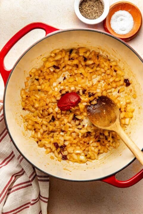 A red enameled Dutch oven containing sautéed chopped onions, a dollop of tomato paste, and a wooden spoon, with small bowls of salt and pepper nearby.
