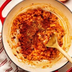 A wooden spoon stirs sautéed diced onions and spices in a red Dutch oven, with small bowls of salt and pepper nearby.