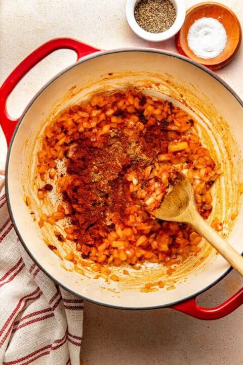 A wooden spoon stirs sautéed diced onions and spices in a red Dutch oven, with small bowls of salt and pepper nearby.