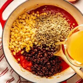 Liquid being poured into a red Dutch oven containing corn, lentils, black beans, and tomato sauce, with wooden bowls of salt and pepper nearby.