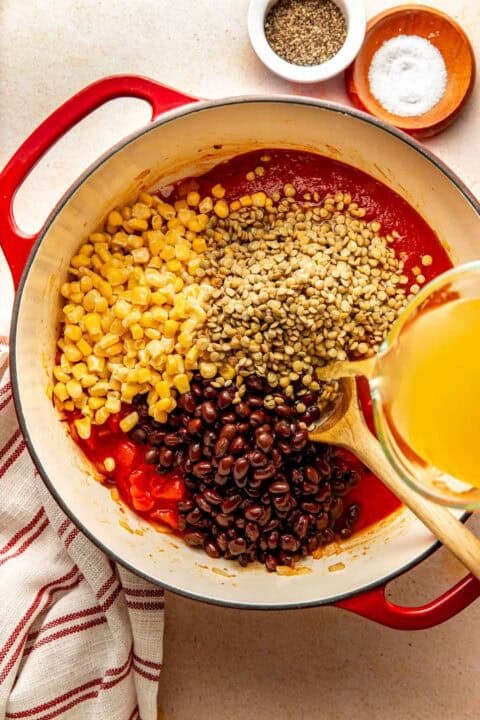 Liquid being poured into a red Dutch oven containing corn, lentils, black beans, and tomato sauce, with wooden bowls of salt and pepper nearby.