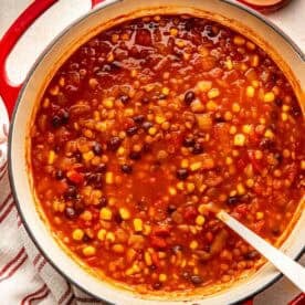 A red Dutch oven filled with a stew of black beans, corn, and tomatoes, with small bowls of salt and pepper nearby.
