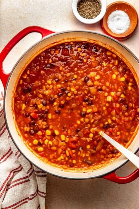A red Dutch oven filled with a stew of black beans, corn, and tomatoes, with small bowls of salt and pepper nearby.