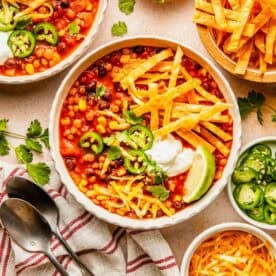 Two bowls of bean and corn tortilla soup garnished with sour cream, jalapeños, cheese, and lime, with side dishes of tortilla strips, sliced jalapeños, and shredded cheese.