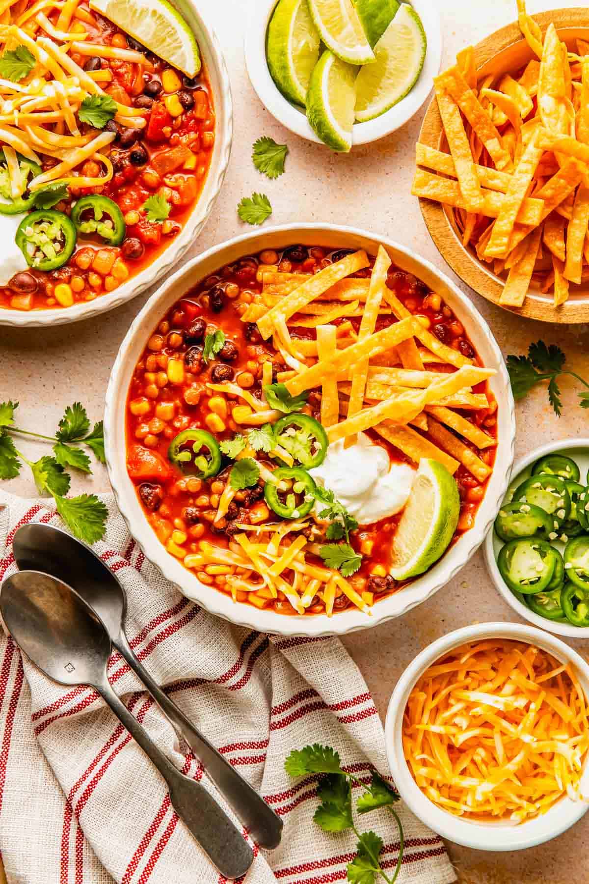 Two bowls of bean and corn tortilla soup garnished with sour cream, jalapeños, cheese, and lime, with side dishes of tortilla strips, sliced jalapeños, and shredded cheese.