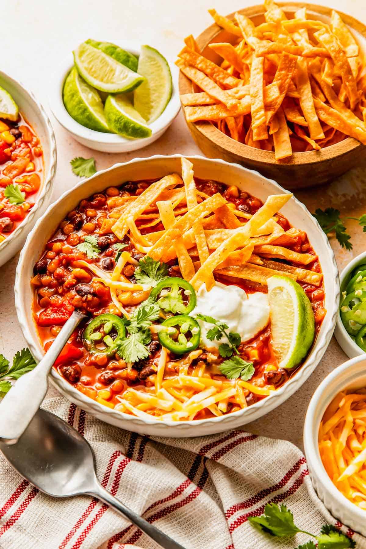 A bowl of bean and corn chili topped with tortilla strips, sour cream, jalapeño slices, and cilantro, served with lime wedges and additional tortilla strips.