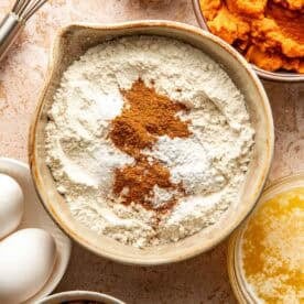 A bowl of flour topped with cinnamon and baking powder, surrounded by bowls of pumpkin purée, brown sugar, melted butter, eggs, and a whisk.