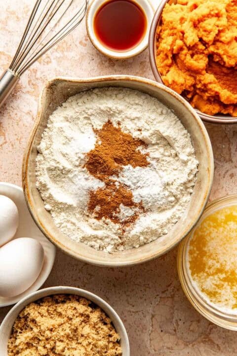 A bowl of flour topped with cinnamon and baking powder, surrounded by bowls of pumpkin purée, brown sugar, melted butter, eggs, and a whisk.