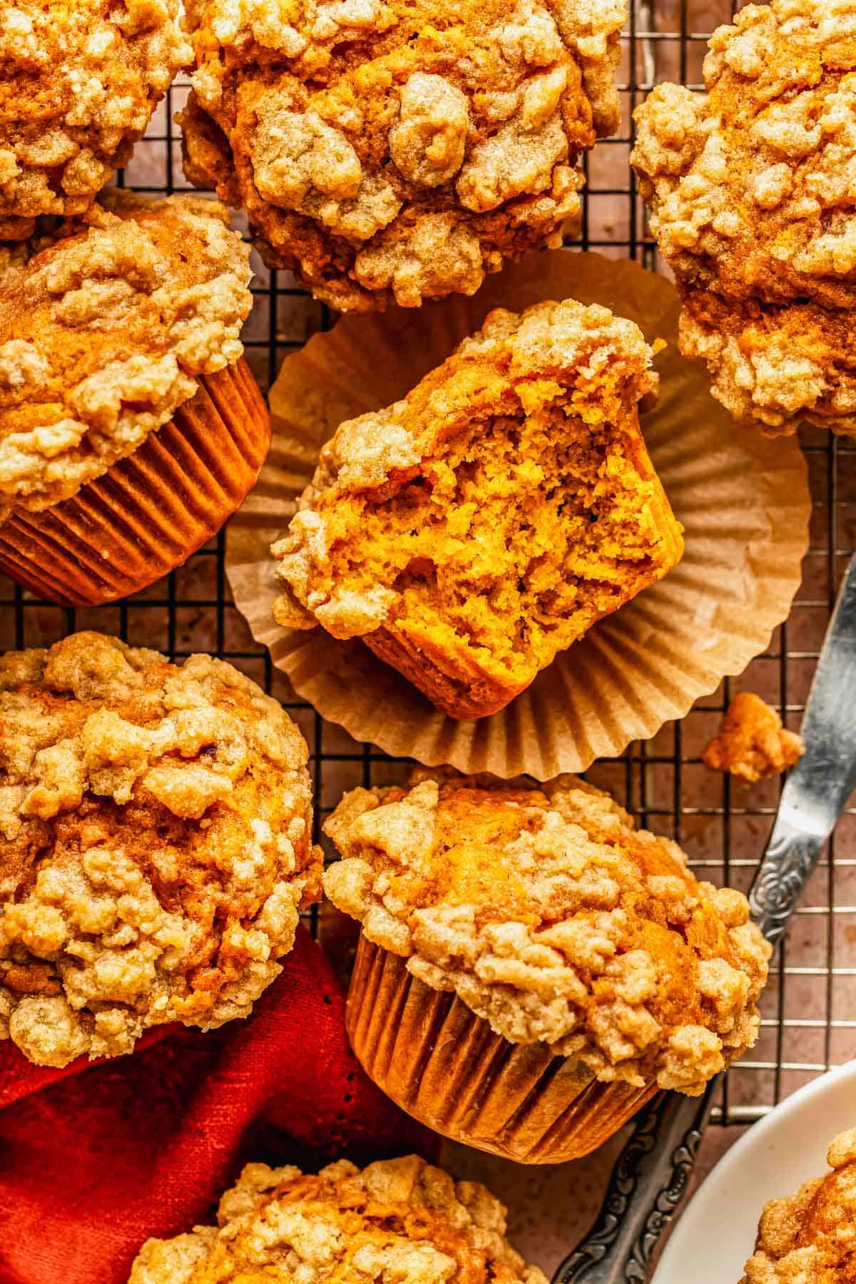 Several pumpkin muffins with streusel topping on a wire cooling rack, one shown broken open to reveal the orange interior.