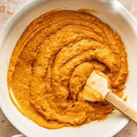 Orange cake batter in a white mixing bowl with a spatula, next to empty eggshells.