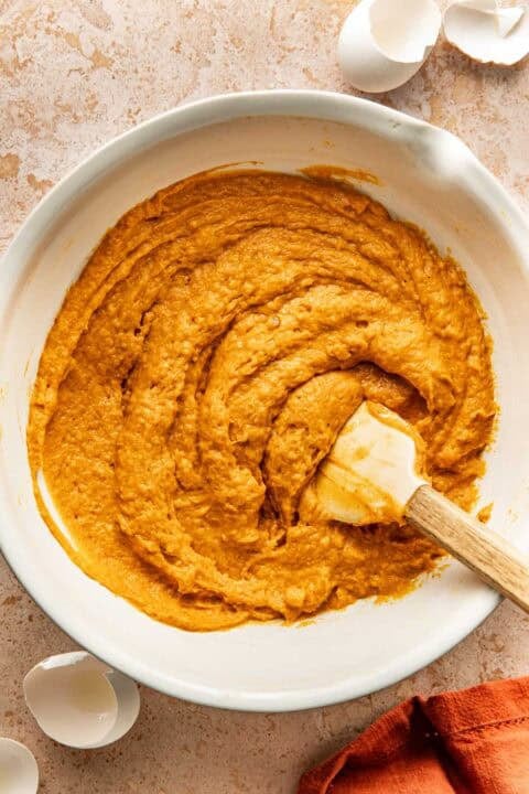 Orange cake batter in a white mixing bowl with a spatula, next to empty eggshells.