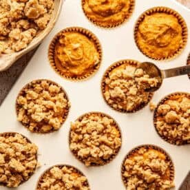 A spoon adds crumb topping to pumpkin muffin batter in a cupcake tin, with a bowl of extra topping nearby.