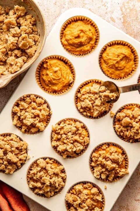 A spoon adds crumb topping to pumpkin muffin batter in a cupcake tin, with a bowl of extra topping nearby.